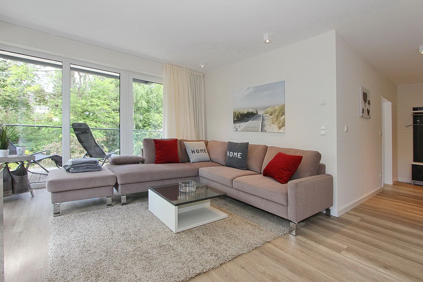 A modern living room with a beige sofa, two armchairs, a glass coffee table, and a view of greenery through large windows.