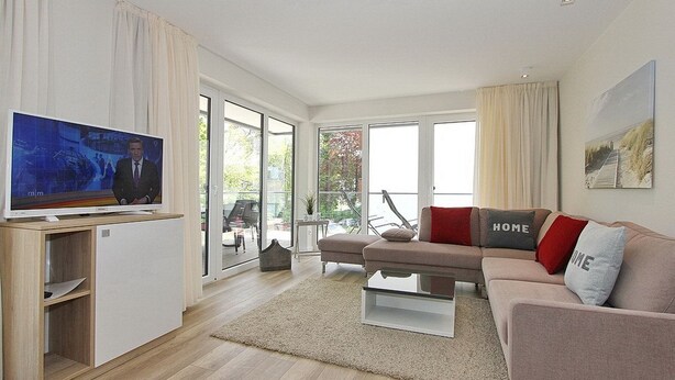 A modern living room with a flat-screen TV, a sofa with 'HOME' pillows, a glass coffee table, and a view through large sliding doors to an outdoor area. - Villa Stern Apartment 10 - Villa Stern (Timmendorfer Strand)