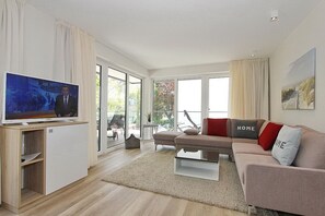 A modern living room with a flat-screen TV, a sofa with 'HOME' pillows, a glass coffee table, and a view through large sliding doors to an outdoor area. - Villa Stern Apartment 10 - Villa Stern (Timmendorfer Strand)