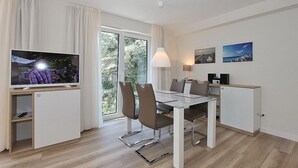 A modern hotel room with a glass dining table, comfortable seating, a flat-screen TV, a desk with a lamp, and a scenic view through the window. - Villa Stern Apartment 11 - Villa Stern (Timmendorfer Strand)
