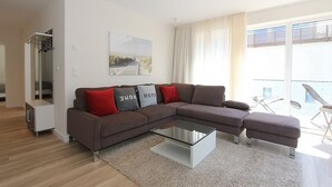A modern living room with a brown sectional sofa, a glass coffee table, and a view of the outdoors through large windows. - Villa Stern Apartment 11 - Villa Stern (Timmendorfer Strand)