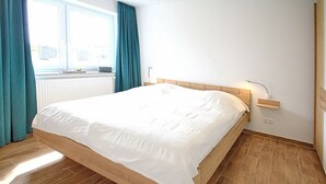 A neatly made bed in a room with wooden flooring, a window with curtains, and a bedside table with a lamp.