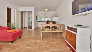 A modern living room with a red sofa, a wooden dining table with benches, a TV mounted on the wall, and a kitchen in the background.