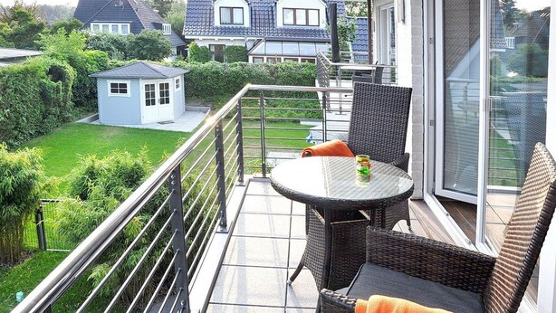 A balcony with wicker furniture, a glass table, and a view of a garden and neighboring houses.
