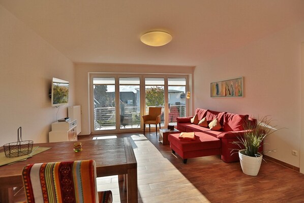 A spacious living room with a red sofa, a dining table, and a large sliding glass door leading to a balcony.