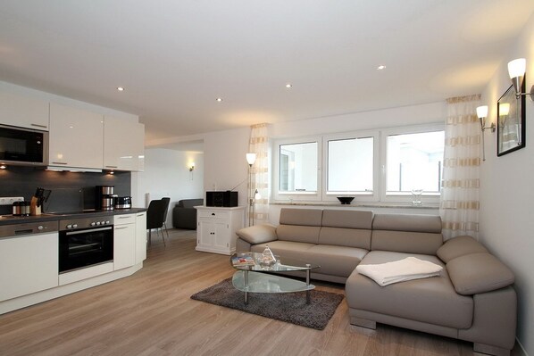 A modern apartment with a kitchen, dining area, and living room. It includes a sofa, a glass coffee table, and a TV.