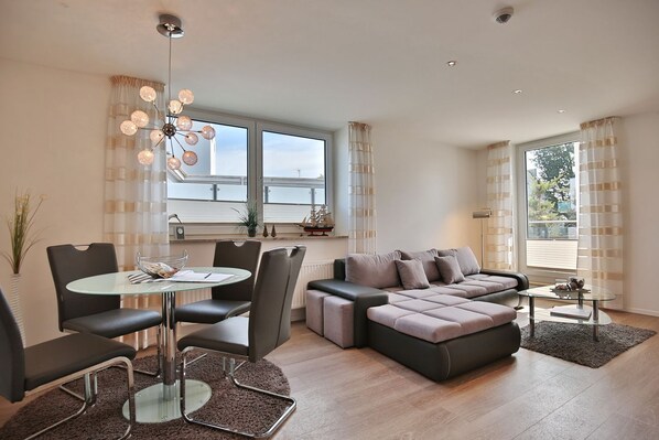 A modern living room with a glass dining table, a grey sofa, and a large window with curtains.