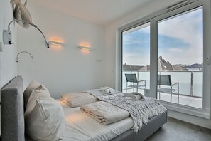 Un dormitorio moderno con un ventanal grande, una cama con ropa de cama blanca, un banco gris y dos luces fijadas a la pared.