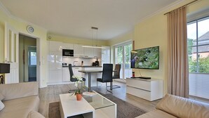 A modern living room with a sofa, coffee table, and TV. There is a kitchen area with white cabinets and a dining table with chairs. - Baltic Sea Apartment 1 - Baltic Sea (Timmendorfer Strand)
