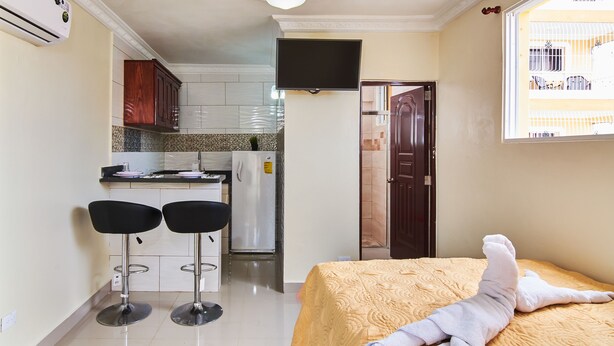 Een compacte hotelkamer met een bed, een kleine kitchenette, twee barkrukken en een aan de muur bevestigde televisie.