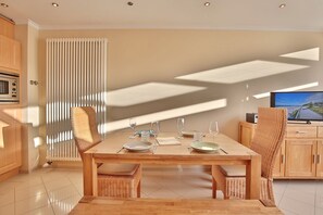 Un'area pranzo moderna con un tavolo di legno apparecchiato per quattro persone, sedie in vimini, una grande finestra con tende a persiana verticale e un televisore su un mobile in legno.