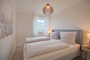 A bedroom with a bed, white bedding, a grey headboard, a bedside table with a lamp, and a hanging light fixture. - Residenz Alte Bergstrasse Apartment 17 - Residenz Alte Bergstrasse (Scharbeutz)