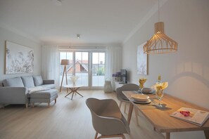 A modern living room with a grey sofa, a dining table set for two, and a large window with a view of buildings. - Residenz Alte Bergstrasse Apartment 17 - Residenz Alte Bergstrasse (Scharbeutz)