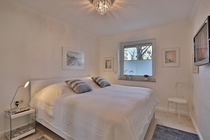 Une chambre avec un grand lit, une table de chevet avec une lampe, une fenêtre avec des stores et une chaise.