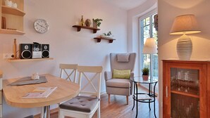 A cozy living room with a round wooden table, two white chairs, a grey armchair, a small side table with a lamp, and a glass cabinet with wine glasses.