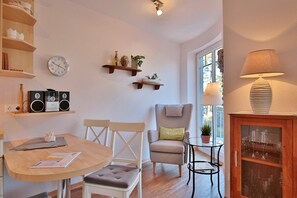 Una sala de estar acogedora con una mesa redonda de madera, dos sillas blancas, un sillón gris, una pequeña mesita de noche con una lámpara y un aparador de vidrio con copas de vino.