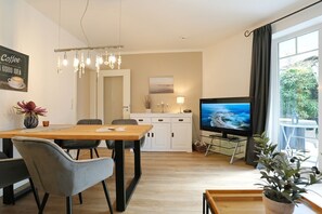 A modern living room with a wooden dining table, two grey chairs, a TV, and a balcony with plants.