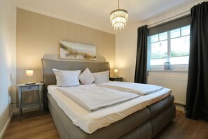 A double bed with a headboard, bedside tables with lamps, a chandelier, a painting on the wall, and a window with curtains.