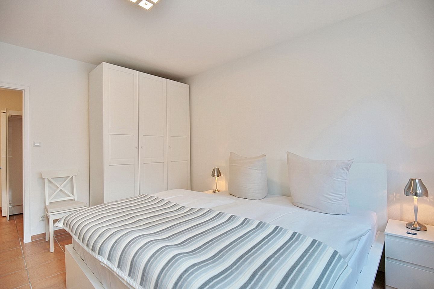 Un dormitorio con cama, armario blanco, lámparas de noche y una silla.