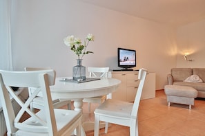 A modern living room with a round dining table, white chairs, a beige sofa, and a television on a stand.