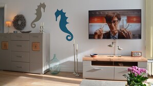 Una sala de estar con televisor, cómoda, jarra con flores y decoración en la pared.