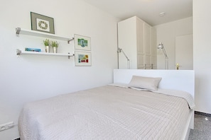 A bedroom with a bed, a nightstand, a shelf with books and a plant, a mirror, and a wardrobe.