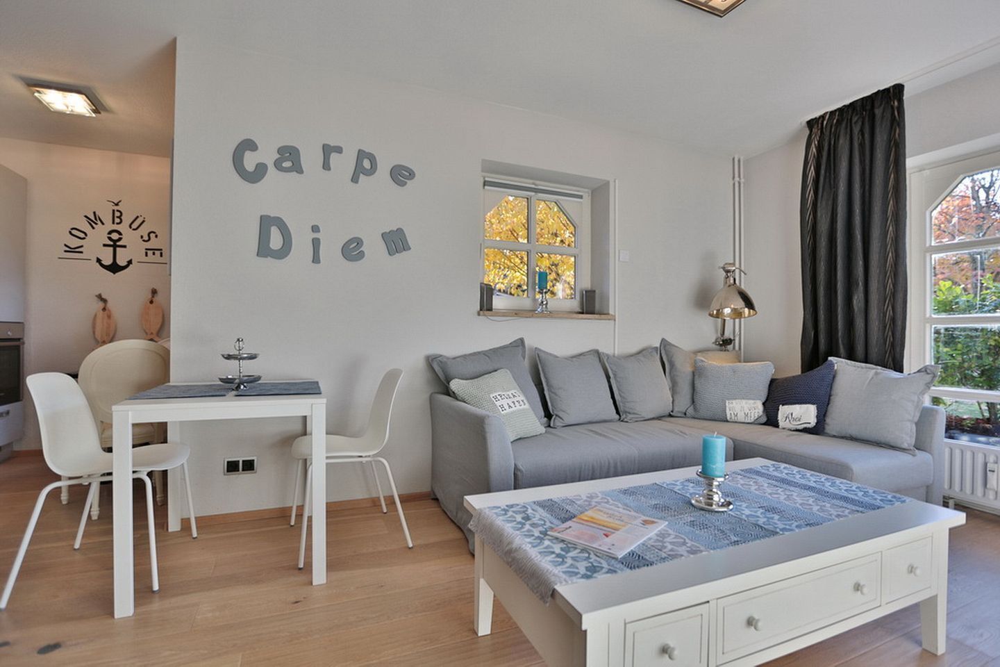 Un saló modern amb un sofà gris, cadires blanques, una taula de menjador i una taula de centre. Es veu una finestra amb fulles d'autorriu, i a la paret hi ha un ancla decorativa i les paraules "Carpe Diem".