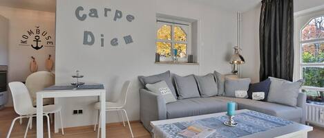 Un saló modern amb un sofà gris, cadires blanques, una taula de menjador i una taula de centre. Es veu una finestra amb fulles d'autorriu, i a la paret hi ha un ancla decorativa i les paraules "Carpe Diem".