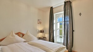 Una cama bien hecha con sábanas blancas en una habitación con piso de madera, una mesita de noche con lámpara y una ventana con cortinas.