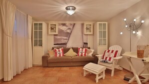 Ein gemütliches Wohnzimmer mit braunem Sofa, Sesseln mit Streifenmuster und einem kleinen weißen Tisch. Es gibt zwei Schränke mit Glastüren, eine Chandelüster und ein großes Fenster mit Vorhängen.