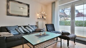 Ein Wohnzimmer mit Glastisch, einer schwarzen Couch mit gestreiften Kissen, einem schwarzen Sessel und einem großen Spiegel an der Wand.
