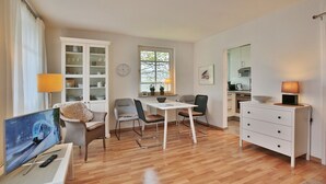 Ein modernes Wohnzimmer mit Holzboden, einem Essbereich mit Tisch und Stühlen, einem weißen Schrank, einem Fernseher und einer Küchenzeile mit Herd und Schränken.