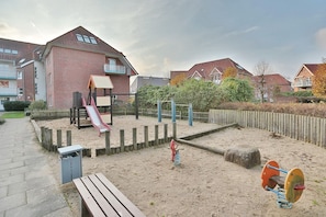 A playground with a slide and climbing structures, surrounded by sand and wooden fences.