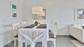 Un coin repas avec une table dressée pour quatre personnes, des chaises blanches, un lustre et des tableaux encadrés accrochés aux murs.