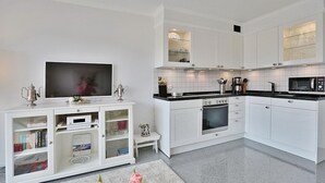Une cuisine moderne avec des meubles blancs, un four encastré, un four à micro-ondes et un téléviseur posé sur une console blanche.