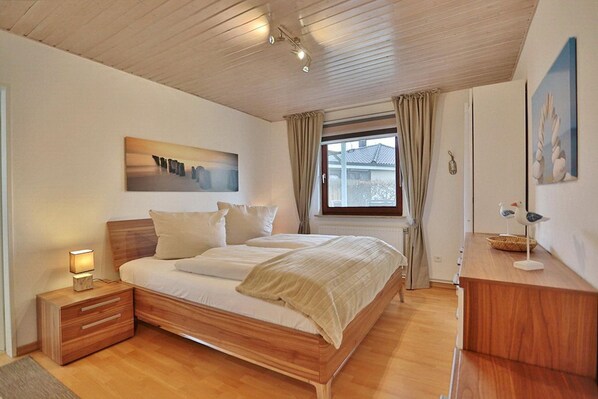 A bedroom with a wooden bed, a nightstand with a lamp, a window with curtains, and a painting on the wall.