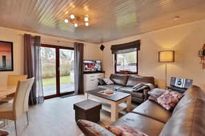 A modern living room with a sofa, coffee table, and TV. There is a dining area with a table and chairs. The room has a large window and a ceiling with a light fixture.