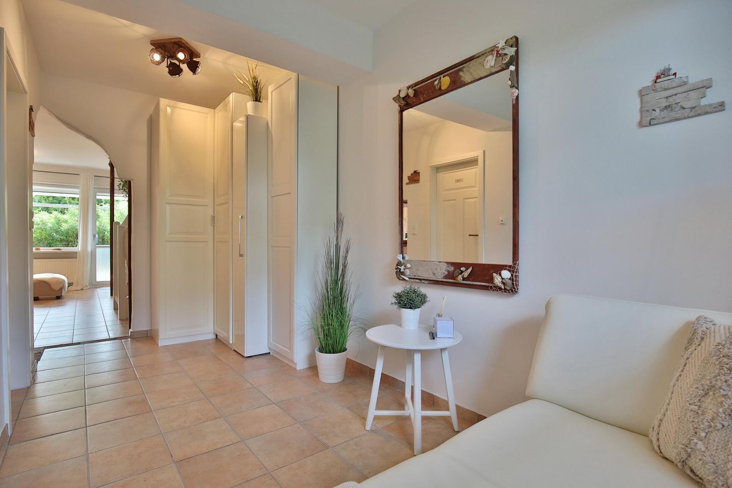 Un pasillo con un sofá blanco, una mesa redonda con una planta en maceta, un espejo y una puerta que da a otra habitación.