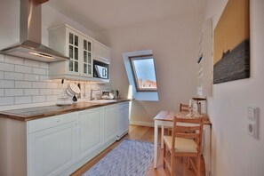 Cocina compacta con muebles blancos, horno empotrado y ventana con vista al cielo.