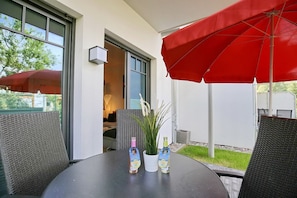 Un patio con sillas de mimbre, un paraguas rojo y dos botellas de agua sobre una mesa.
