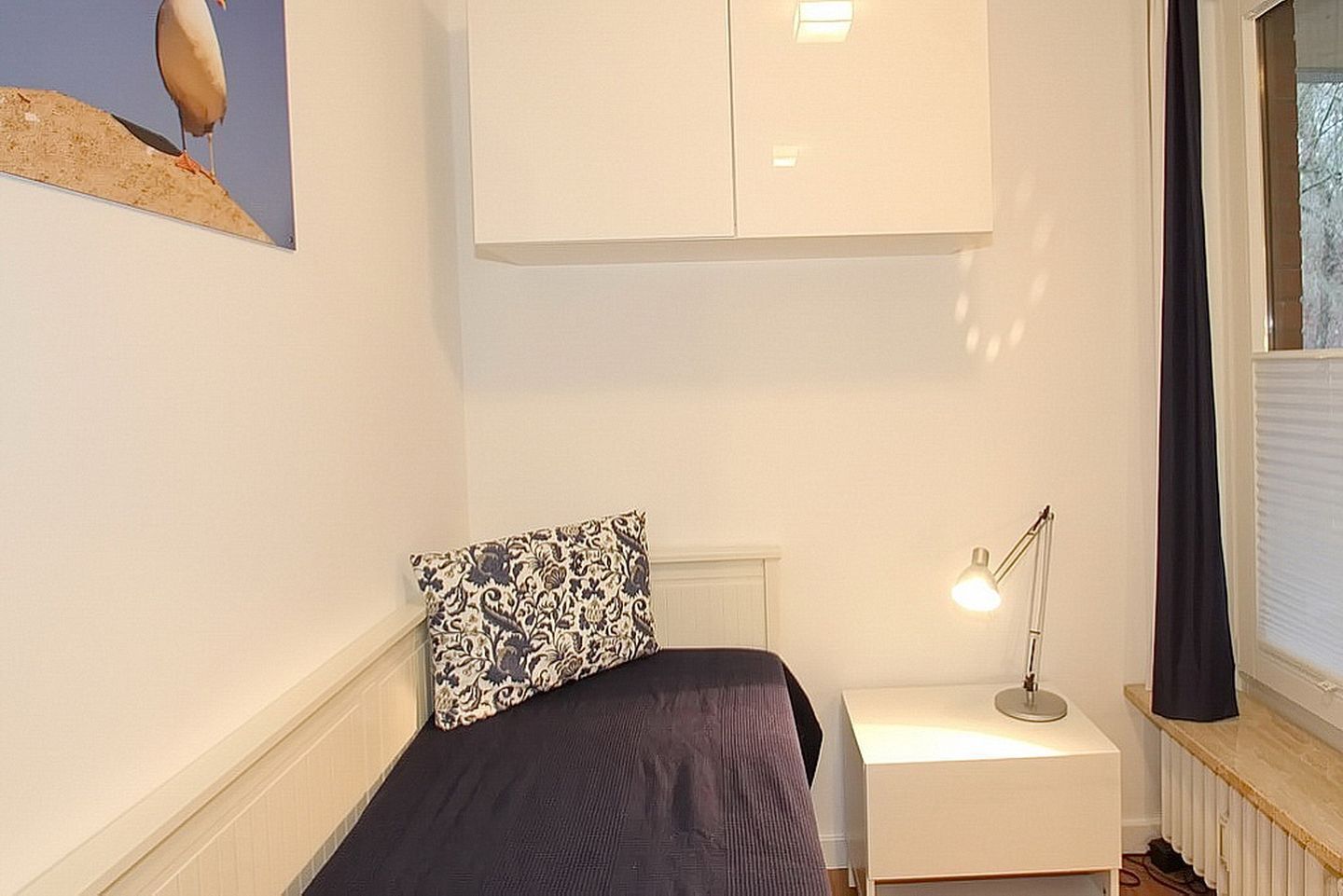 A bedroom with a bed, a nightstand, a lamp, a window with curtains, and a wall-mounted cabinet.