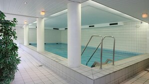 An indoor swimming pool with tiled walls and a tiled floor, surrounded by white pillars and a potted plant.