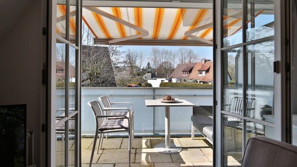Ein Balkon mit Glastür, einem Tisch, zwei Stühlen und einem gestreiften Sonnensegel.