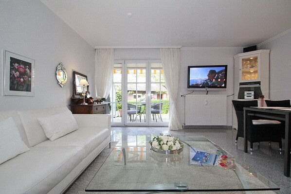 A modern living room with a white sofa, a glass coffee table, a flat-screen TV, and a dining area with a table and chairs.