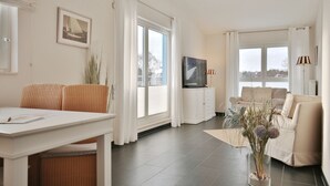 A modern living room with a dining area, large windows, and a flat-screen TV on a white cabinet. - Penthouse am Platz Apartment 10 - Penthouse am Platz (Timmendorfer Strand)