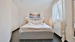 A bedroom with a bed, white bedding, a painting above the bed, and a window with curtains. - Penthouse am Platz Apartment 10 - Penthouse am Platz (Timmendorfer Strand)