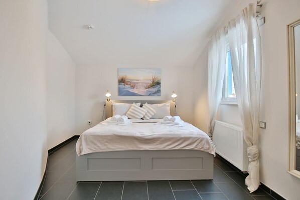 A bedroom with a bed, white bedding, a painting above the bed, and a window with curtains.