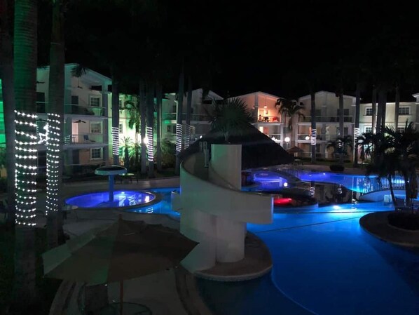 A nighttime resort pool area with illuminated water features and a central structure.