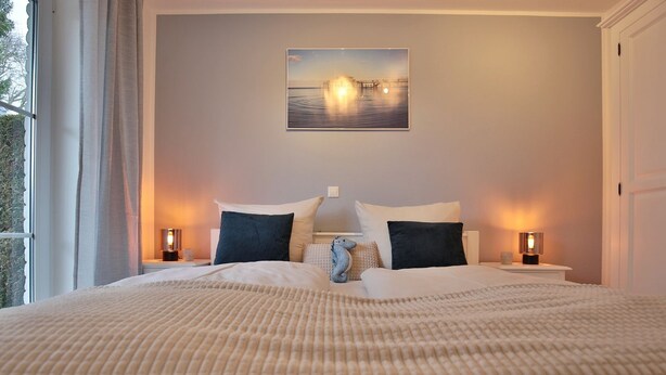 Un lit bien fait, recouvert d’une couverture beige à texture intéressante, avec deux oreillers et un petit ours en peluche. Il y a deux tables de chevet avec des lampes et, au mur, un tableau représentant un pont.