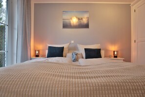 A neatly made bed with a textured beige blanket, two pillows, and a small teddy bear. There are two bedside tables with lamps and a framed picture of a bridge on the wall.